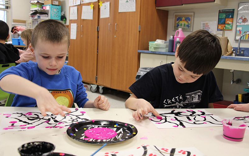 Two first grade students, one with very short hair wearing a blue shirt and the other with dark hair wearing a black tshirt, put pink blossoms on a painting using their thumbs dipped in pink paint.