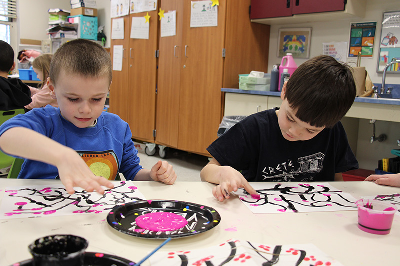 Two first grade students, one with very short hair wearing a blue shirt and the other with dark hair wearing a black tshirt, put pink blossoms on a painting using their thumbs dipped in pink paint.