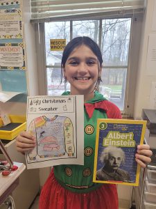 An elementary-age girl smiles broadly as she holds up a book about Albert Einstein and a page she colored.