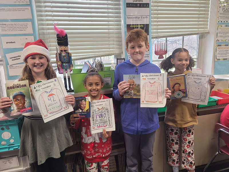 A group of four fifth-grade students each hold up a book they read and a page they colored, creating an "ugly" sweater  based on the accomplishments of the person they read about. There are two girls on the left, then a boy and another girl. All are smiling.