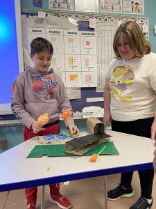 Two fourth-grade students stand at a table showing the environment they built. It includes a green patch of material and things used by native Americans in New York.