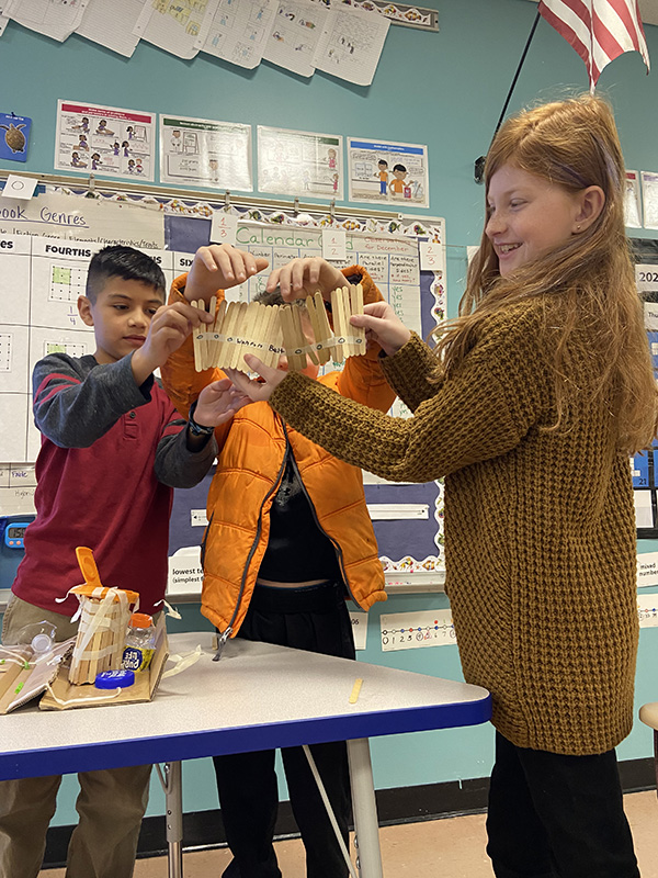 A group of three students hold up a structure made out of wooden popsicle sticks.