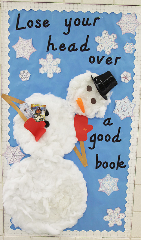 A bulletin board with a light blue background that says Lose your head over a good book. There are large snowflakes and a large snowman whose head is tipping off.