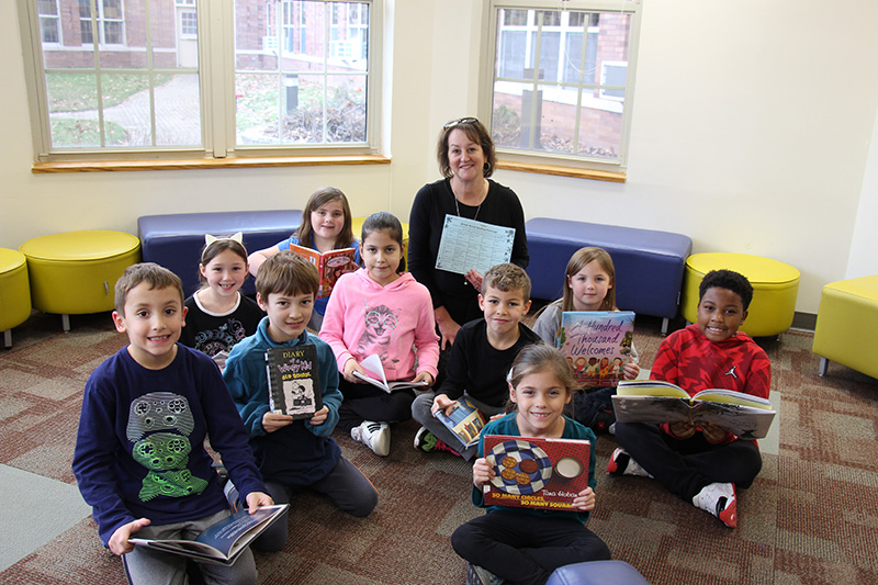 Nine second-grade students sit on a floor, each holding a book. In the back is a teacher with shin-length brown hair smiling with them.