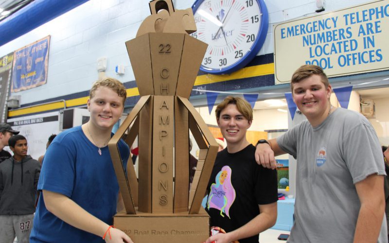 Three high school boys hold a large trophy made of cardboard that says 22 Champions. 2022 Boat Race Champions. They are all smiling.