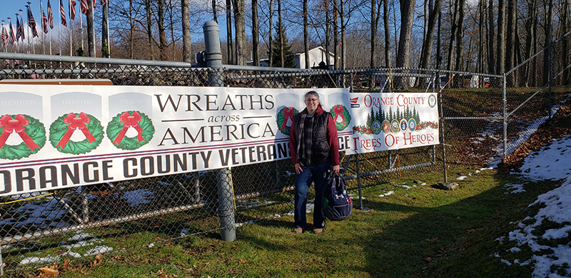 A woman wearing blue jeans, a plaid scarf, red hoodie and black vest stands in front of a sign that says Wreaths Across America. 