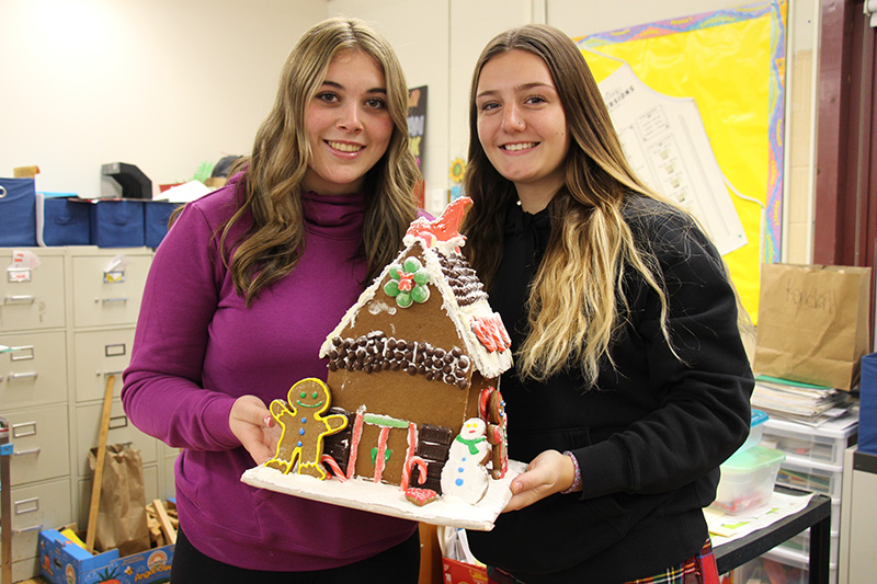 Two high school age young women smile and hold a large gingerbread house they made. They both have long hair. The girl on the left is wearing a purple turtle neck shirt and the girl on the right is wearing a black sweater. The gingerbread house is decorated with cookies, multi-colored candies and chocolate chips.
