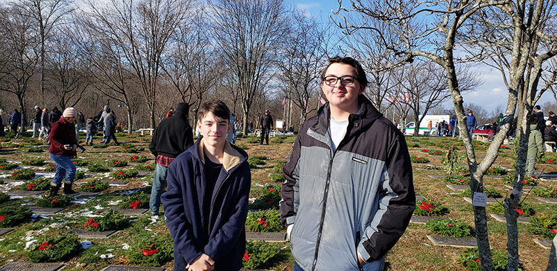 Two high school students stand in the cemetery. The student on the left has short hair and is wearing  a blue zipped jacket. The studen ton the right is taller, has glasses and short hair, is smiling and is wearing a black and gray zipped jacket. There are bare trees and a blue sky behind them.