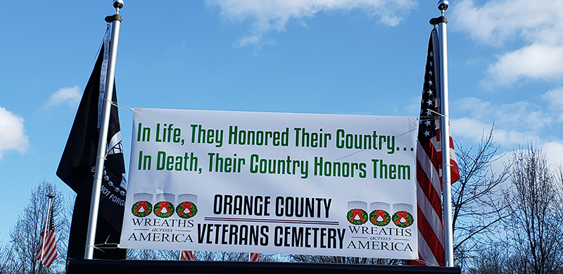 A black prisoner of war flag on the left and an American flag on the right. In the center is a sign that says In life, they honored their country... In death, their country honors the. Orange County Veterans Cemetery. There is blue sky above it.