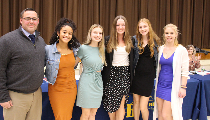 A group of five high school age young women stand arm in arm with their coach, a man, on the left.