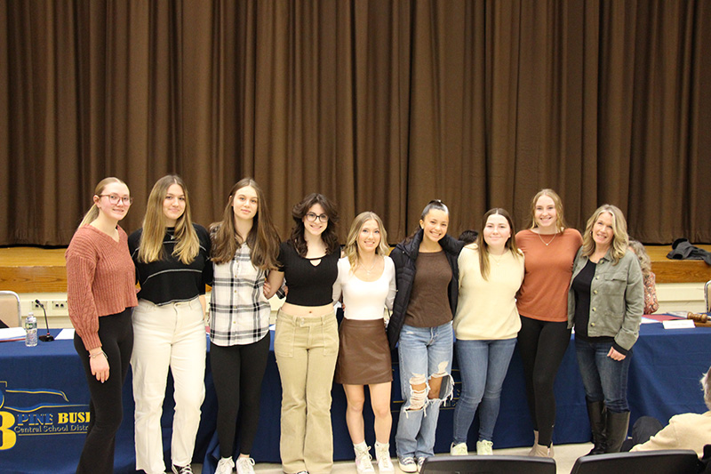 A group of eight high school age women stand arm in arm with their coach, a woman, on the right.