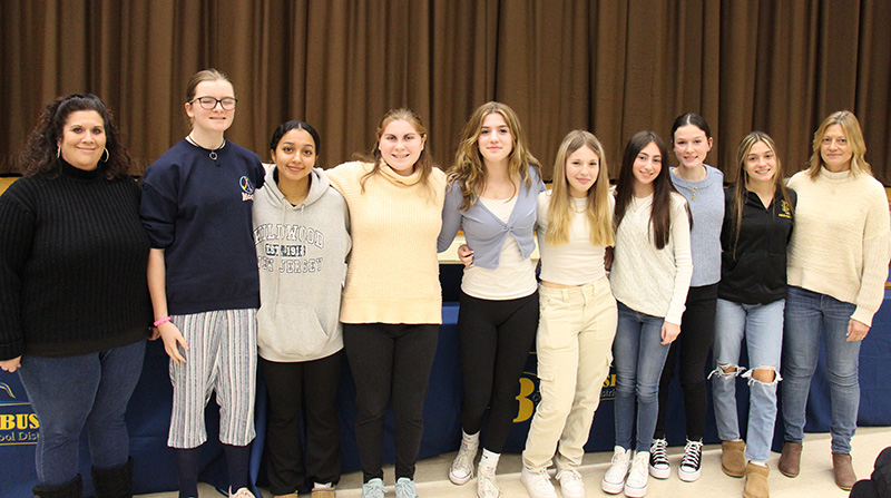 Nine high school age young women stand arm in arm with their coach, a woman, standing with them on the left.