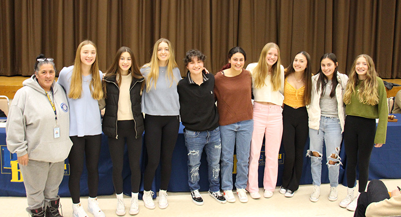 A group of nine high school age girls stand in front of an auditorium. they are arm in arm and their coach, a woman, is standing with them on the left.