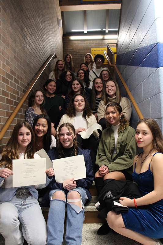 A large group of young women, all high school age, sit on steps going up a to a landing. Some are holding certificates. They are smiling.