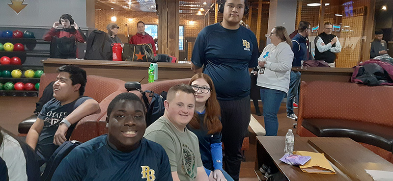 Sitting in a bowling alley, a group of four young men and women sit together smiling.