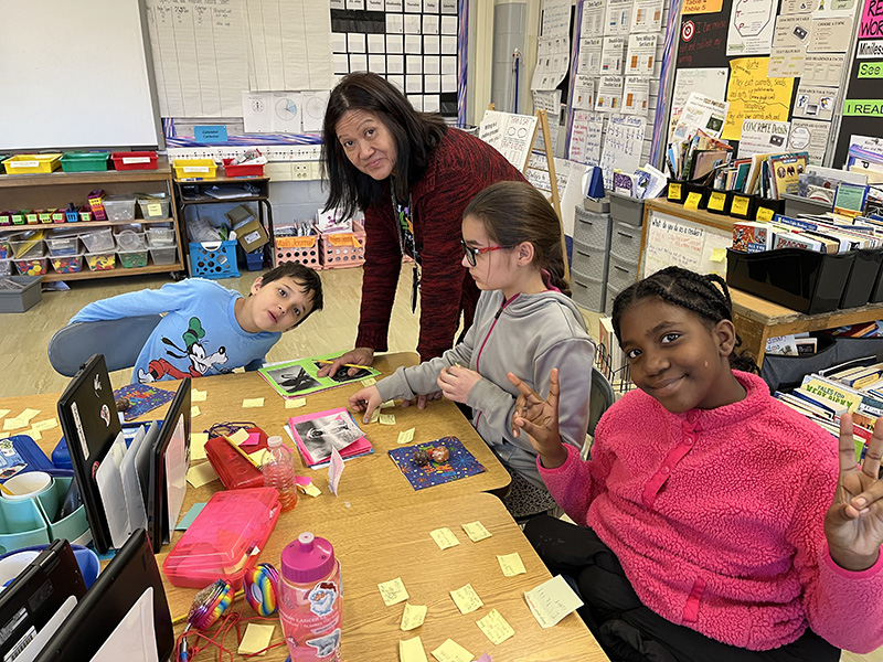 An adult woman with dark, shoulder-length hair, leans onto a table with a group of fourth-grade students who are sitting at it. There is a boy on the right with short dark hair wearing a blue shirt, a girl with her hair pulled back in the center, and a girl with dark hair pulled back smiling, giving peace signs. She is wearing a pink shirt. On the table are books and post-it notes.