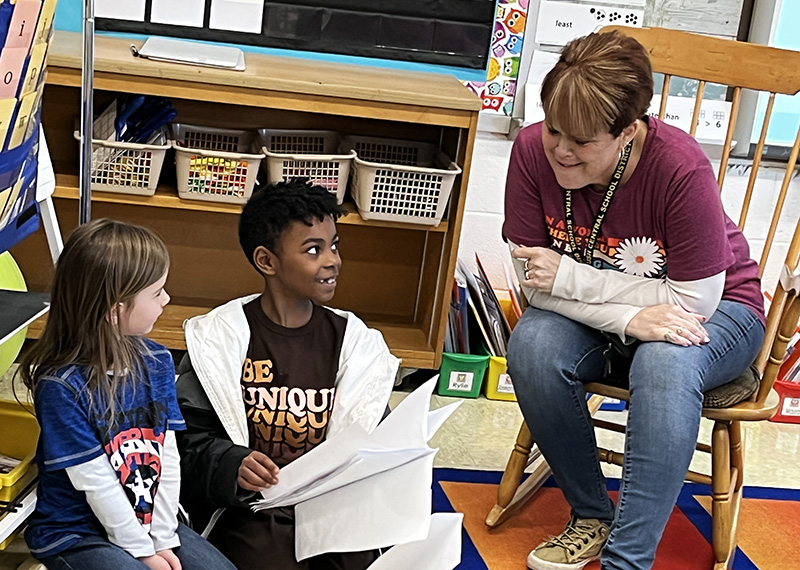 A kindergarten student with long brown hair wearing a blue shirt sits looking at a fourth-grade student with shorter dark hair, wearing a brown shirt that says "Be Unique."  They are both sitting on the floor. The student is looking at a grown up sitting on a chair smiling. They are all smiling.