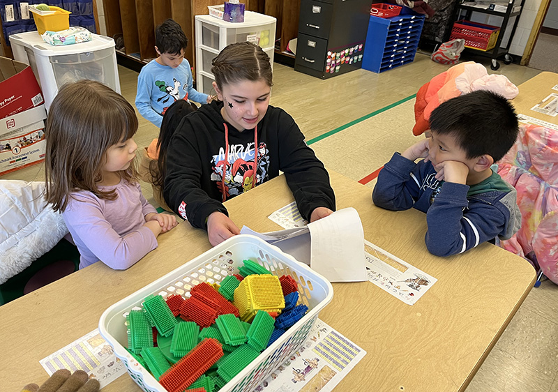 A fourth-grade student sits at a table reading a book to two kindergarten students. On the table is a white basket filled with green, red and yellow blocks.