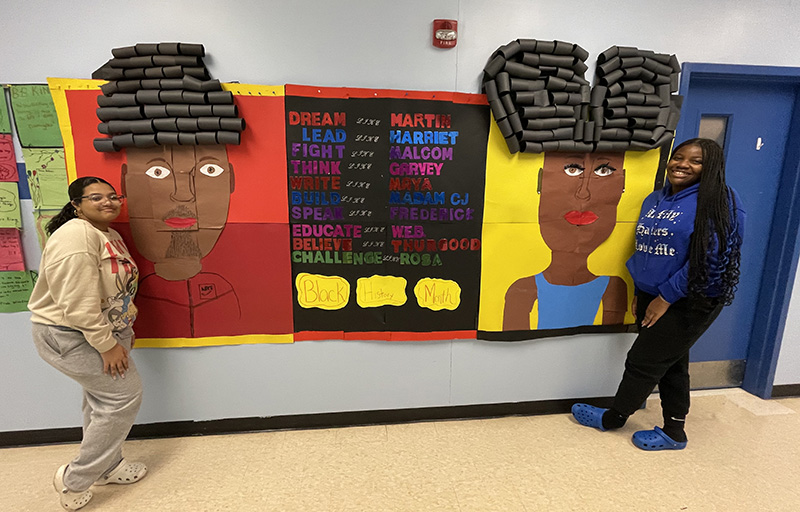 Two middle school girls stand on eithe rside of a large bulletin board that depicts a black man on the left and a black woman on the right. In the center it says Dream like martin, Lead like Harriet, Fight like Malcolm, Think like Garvey, Write like Maya, Build like Madam CJ, Speak like Frederick, Educate like W.E.B., Believe like thurgood, Challenge like Rosa