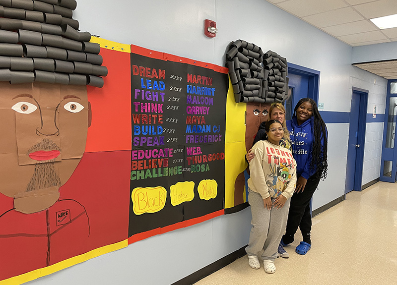 Two middle school girls stand with a woman at the right of a bulletin board that depicts a black man on the left and a black woman on the right. In the center it says Dream like martin, Lead like Harriet, Fight like Malcolm, Think like Garvey, Write like Maya, Build like Madam CJ, Speak like Frederick, Educate like W.E.B., Believe like thurgood, Challenge like Rosa
