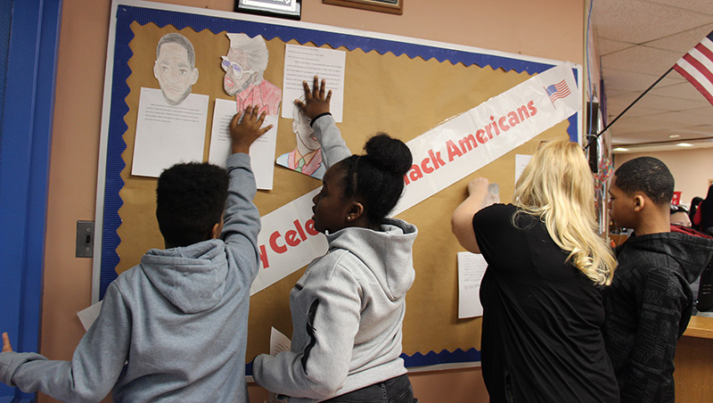 Three students put drawings and biographies on a bulletin board with the help of a woman.