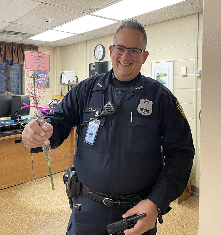 A man in a navy blue police officer's uniform, with short hair and glasses, smiles and holds out a red carnation.