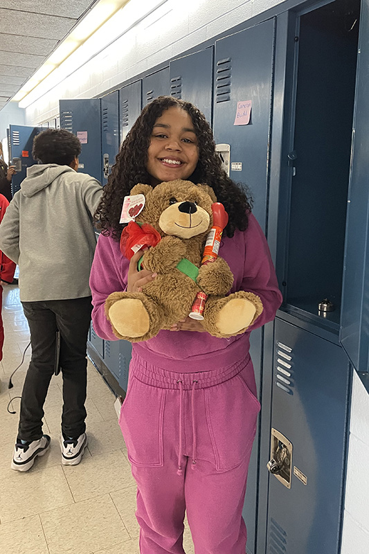 A middle school girl dressed in a mauve sweatshirt and pants, holds a teddy bear valentine.
