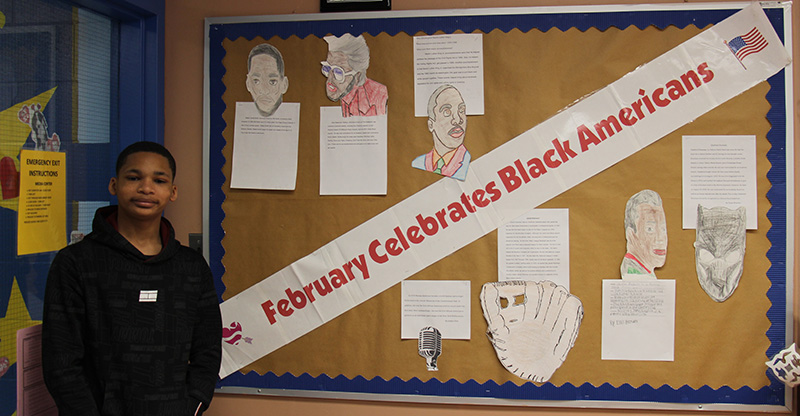 A middle-school boy stands next to a bulletin board that says February Celebrates Black Americans. On the board are pictures and bios of influential Black Americans.