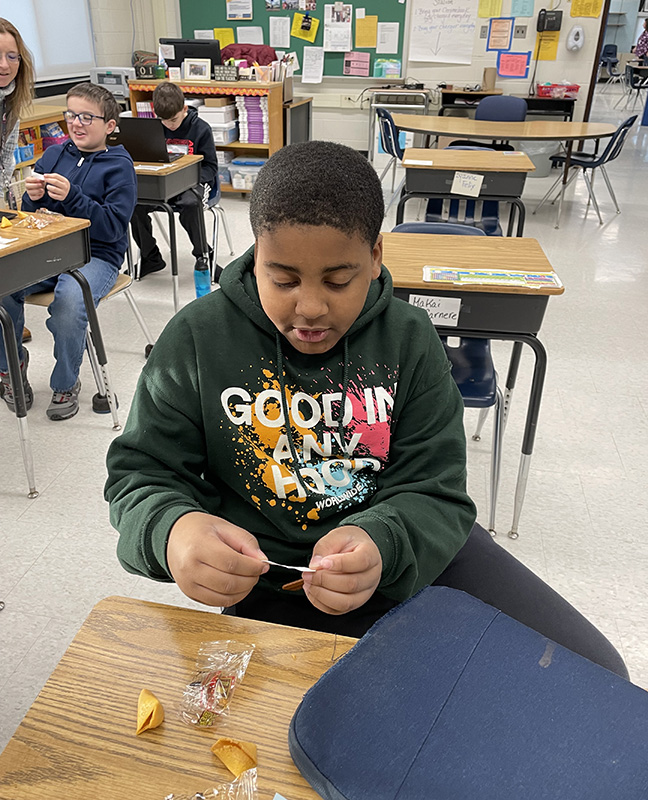 A sixth grade boy with short dark hair, wearing a black hoodie with blue, yellow and pink splashed on the front, sits at a desk reading a fortune after opening a fortune cookie.