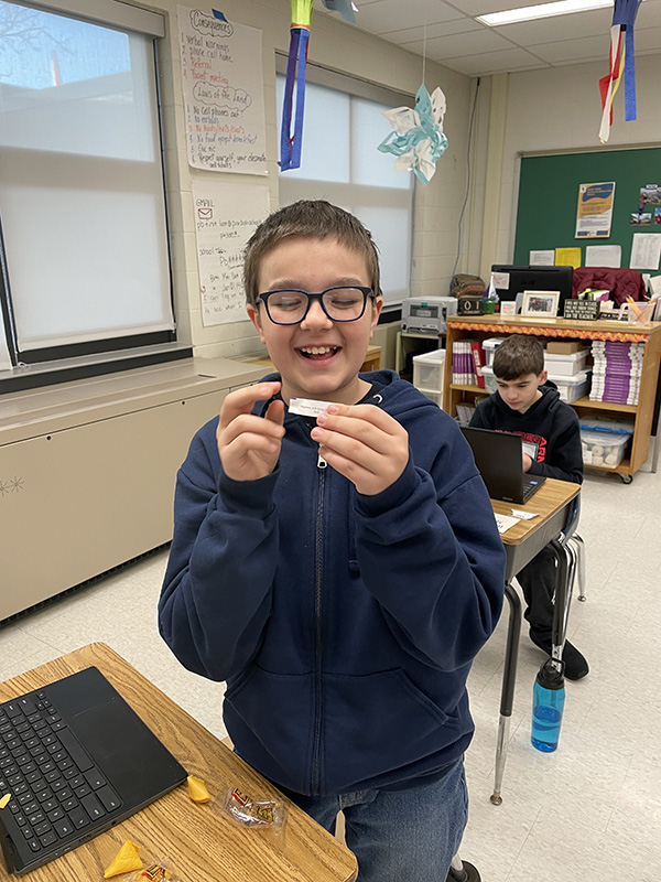 A sixth-grade boy with short brown hair and glasses, wearing a blue hoodie, holds his fortune from a fortune cookie in front of him. He is smiling.