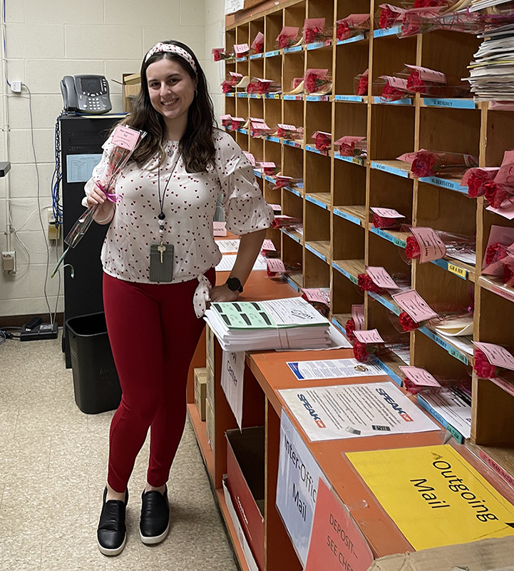 A woman wearing red pants and a red and white shirt holds a red carnation in a clear celophane wrapping. In the mailboxes next to her, there is a red carnation in each box.