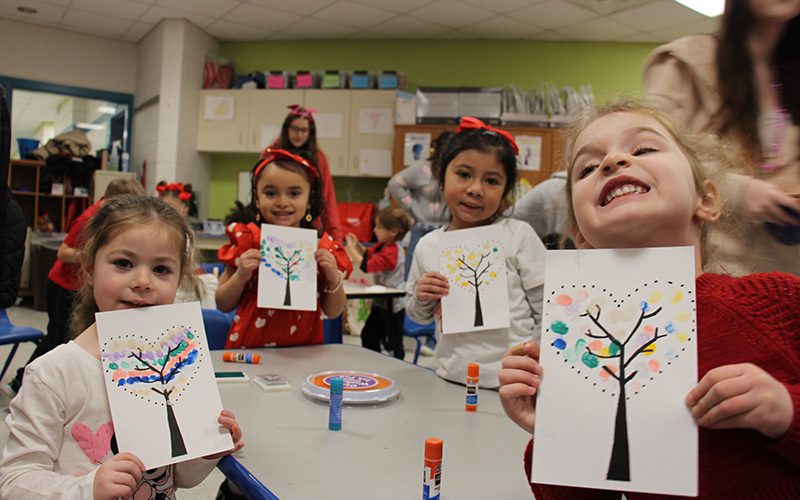 Four pre-K girls smile and hold up their paper with trees they made, with paint with their fingers.