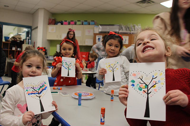 Four pre-K girls smile and hold up their paper with trees they made, with paint with their fingers.