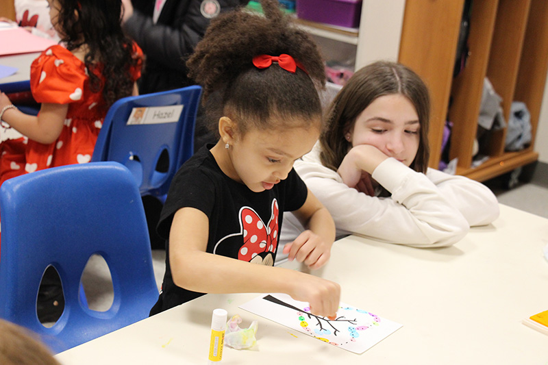 A middle school girl with a white shirt on and long dark hair, squats next to a pre-K  little girl wearing a black tshirt with Minnie Mouse on it. She also has a red ribbon in her hair around her ponytail.