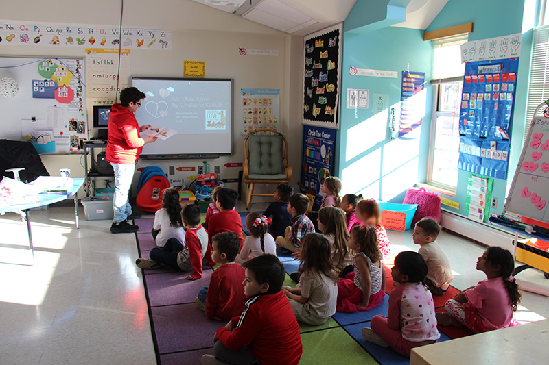 A class filled with pre-K kids sitting on a multi-colored rug, listen as a middle school boy reads to them.