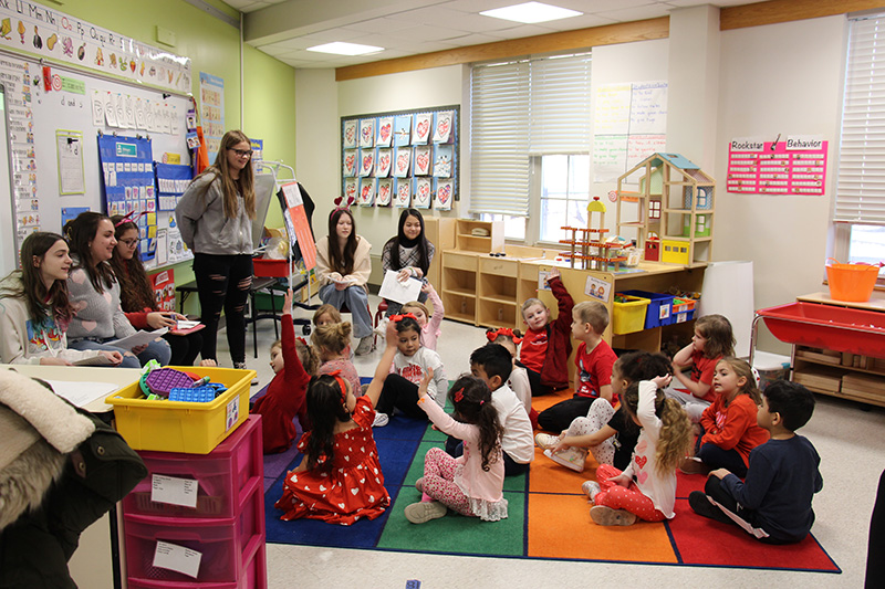 Middle school students stand at the front of a class talking   to a classroom filled with pre-K students sitting on the floor. Some of the students have their hands up.