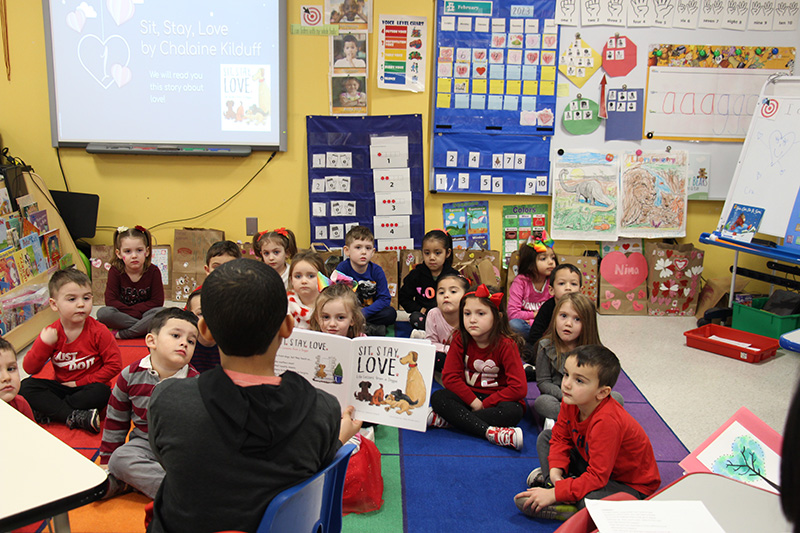 A classroom filled with pre-K students sit on a multi-colored rug as a middle school boy, with dark hair, reads a book to them, showing them the pages.
