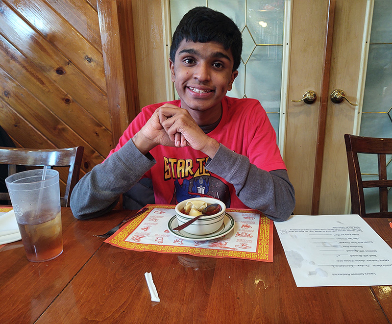 A young man in a red shirt and dark hair smiles. There is soup in front of him.