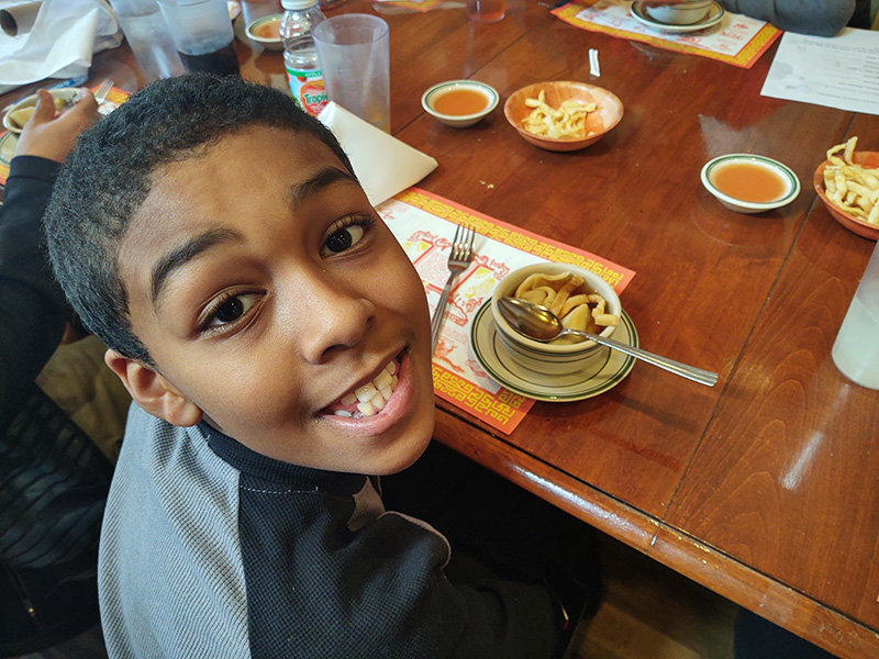A middle school age kid turns toward the camera and smiles big. On the table in front of him is soup.