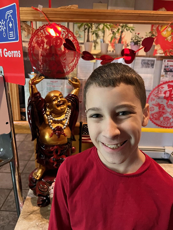 A middle school age boy with short hair wearing a red shirt smiles. There is a Chinese statue behind him.