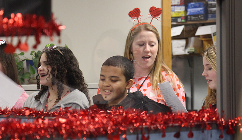 Middle school students and a teacher, with a headband that has hearts coming from it, sing together.