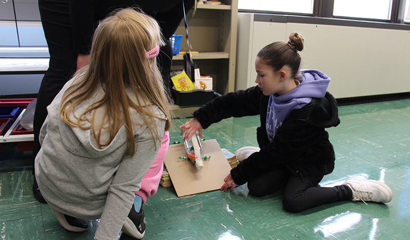 Two fourth-grade girls sit on the floor and put a homemade car on a ramp to see it go.