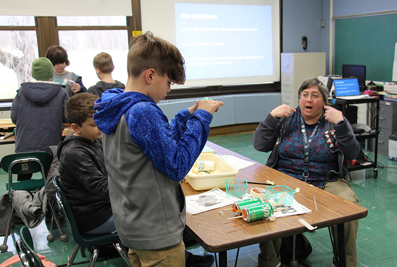 A woman with short gray hair sits at a table with fourth-grade boys who are making a car out of recycled materials. She is talking to them.