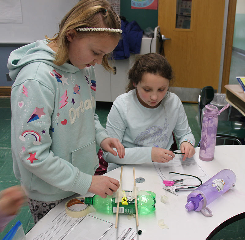 Two fourth-grade girls work on creating a car out of a plastic soda bottle and other recycled items.