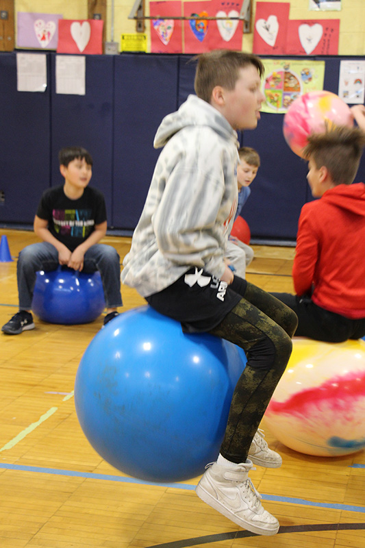 An older elementary age student sits and hops on a large blue hoppity ball.