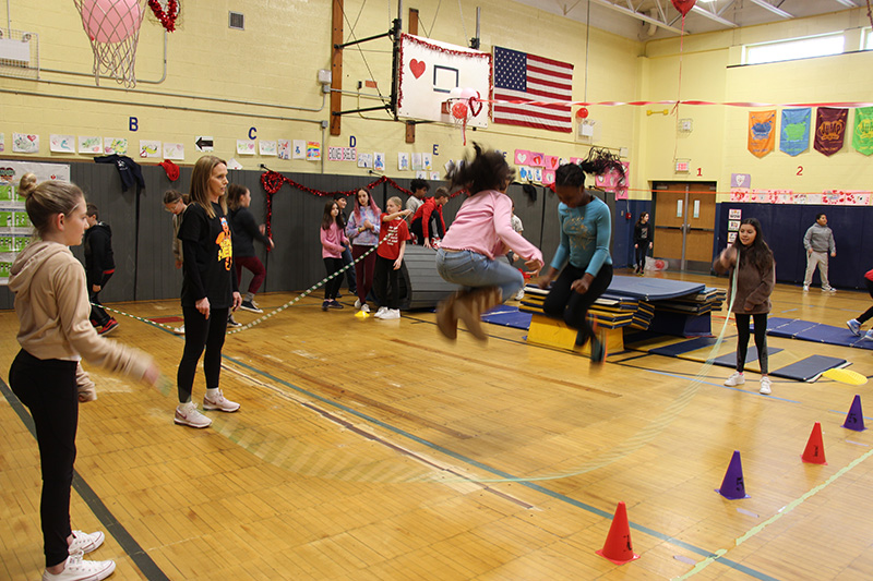 Two elementary girls jump really high as they jump rope with two other kids turning the rope. A woman stands and watches.