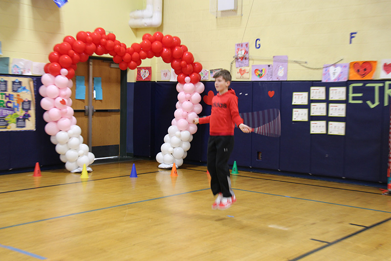 An elementary age boy in a red long-sleeve shirt and black sweatpants jumps rope. There is a red, pink and white heart of balloons behind him.