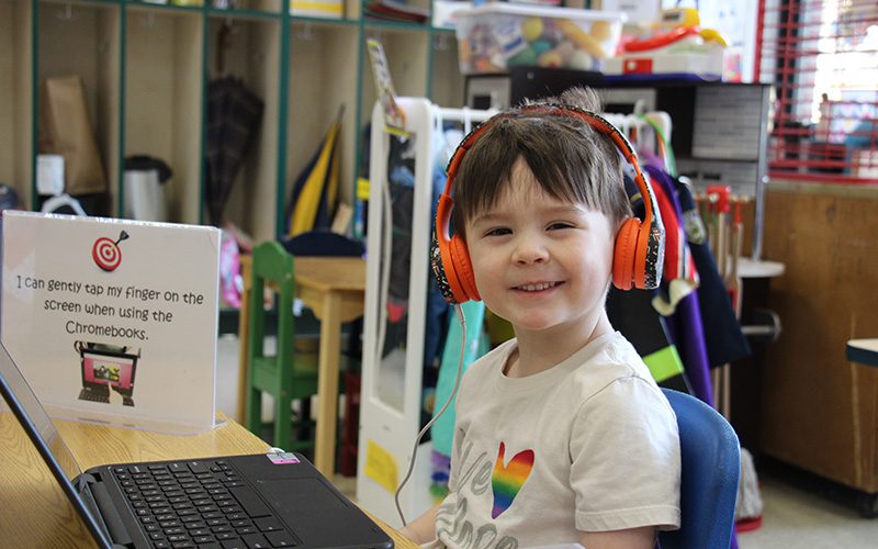 A pre-K kid with short dark hair with big orange headphones on smiles broadly while on a Chromebook.