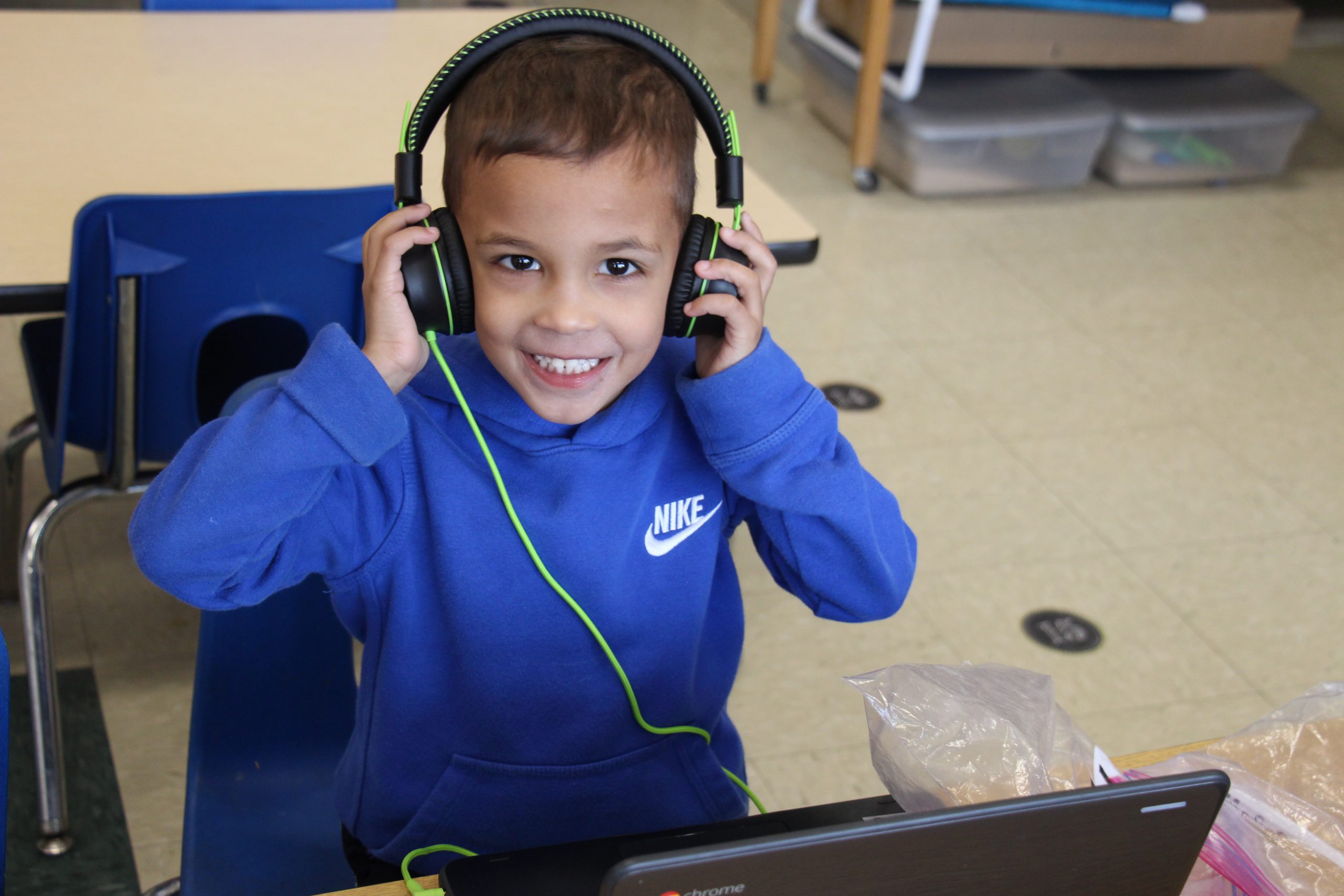 A pre-K boy in a bright blue sweatshirt smiles as he holds his black and green headphones over his ears. He has short dark hair and there is a Chromebook in front of him.