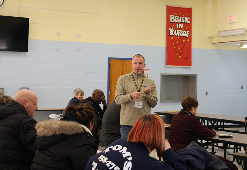 A man in a beige sweater stands in front of a group of adults talking.